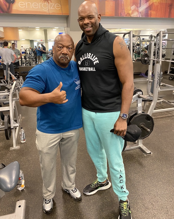 Marc Jackson, former NBA player and now 76ers TV analyst, stands next to his trainer, TK. Jackson is a tall Black man who is bald and smiling and wearing a sleeveless black hoodie with the words BALLISLIFE BASKETBALL and teal sweatpants. The shorter trainer is Black, wearing a serious expression, long mustache, blue t-shirt, light-color pants and sneakers and giving the thumbs up.