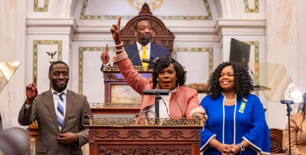 Philadelphia Mayor Cherelle Parker (center) raises her right index finger in the air during her March 14, 2024 budget address. She is a Black woman iwth shoulder-length hair wearing a light red jacket and white blouse, standing behind a podium. Members of City Council around her, left to right: Isaiah Thomas, a Black man standing, wearing an olive suit and blue-and-white-striped tie, partially raises his right index finger. Elevated behind Parker is Council President Kenyatta Johnson, wearing a blue suit and yellow tie, also raising a finger. Councilmember Katherine Gilmore Richardson stands on Parker's other side, a Black woman with long, curly black hair in a blue dress with white trim. Photo by Albert Lee.