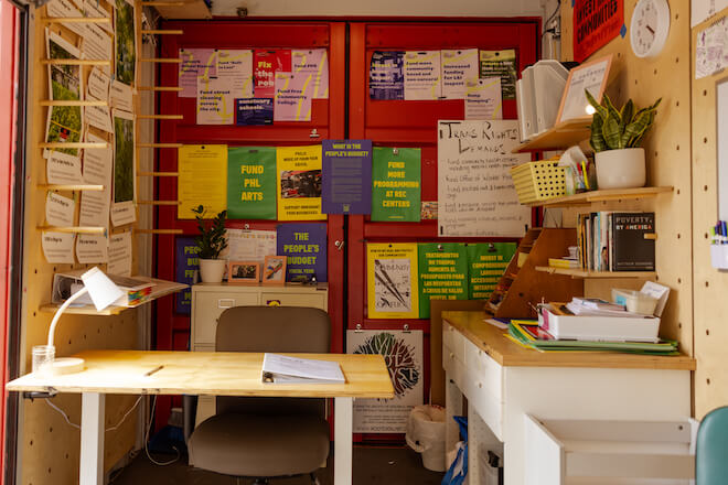Inside the "People's Budget" office in 2023, a red wall with a number of signs requesting things for the city of Philadelphia budget. There's a desk with a lamp in the foreground and shelfs with a plant, paperwork, and a clock.
