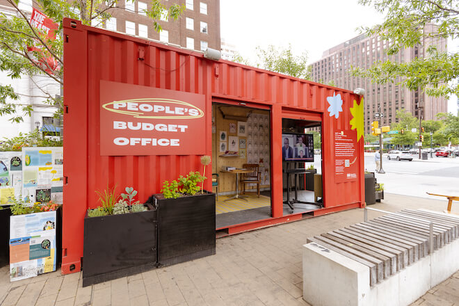 An old shipping container, painted red, stands in LOVE Park in Philadelphia. A sign next to two open door spaces reads "PEOPLE'S BUDGET OFFICE."