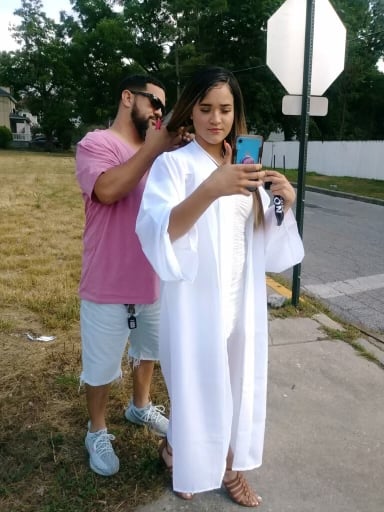 Sahmya Heaven Garcia, a young woman wearing a white graduation robe and holding a cell phone, stands in front of her father, Hector, a Latino man with a pink shirt and shorts who appears to be attaching the clasp of her necklace. They stand on a street corner near a Stop sign.