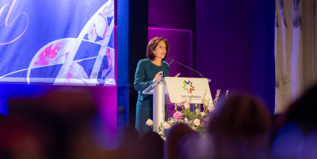 Sue Jacobson, a White woman with shoulder-length brown hair wearing a blue-green dress, stands behind a podium and in front of a purple backdrop to speak at the 2024 Paradigm Awards. Photo courtesy of the Chamber of Commerce of Greater Philadelphia.
