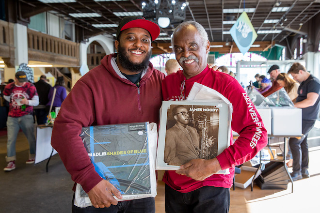 Two Black men in red sweatshirts stand side by side, each holding a vinyl record at VinylCon.