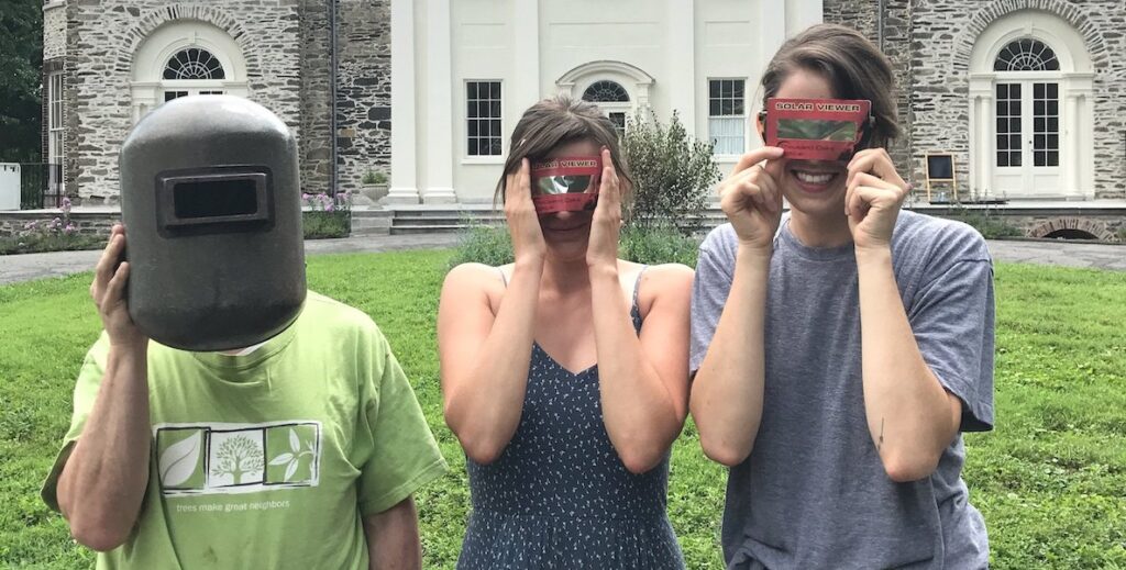 Three white people stand before Hamilton Mansion in The Woodlands in West Philadelphia. Two hold up eclipse viewers to their eyes. The other holds up protective equipment.