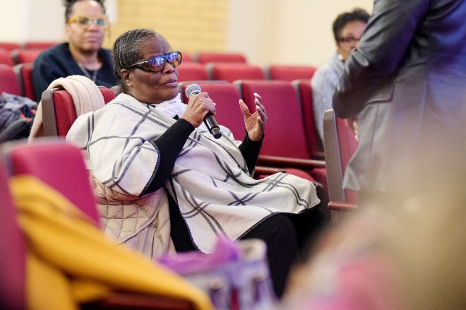 Advocate Yvonne Hughes, an older African American woman, sits in one of many red chairs at a community meeting and holds a microphone. Her hair is close to her head. She is smiling and wearing sunglasses, long earrings and a cream wrap with dark plaid lines. Around her are other people.