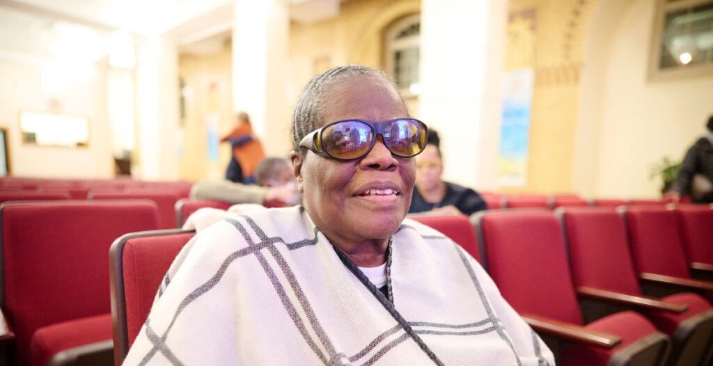 Advocate Yvonne Hughes, an older African American woman, sits in one of many red chairs at a community meeting. Her hair is close to her head. She is smiling and wearing sunglasses, long earrings and a cream wrap with dark plaid lines.