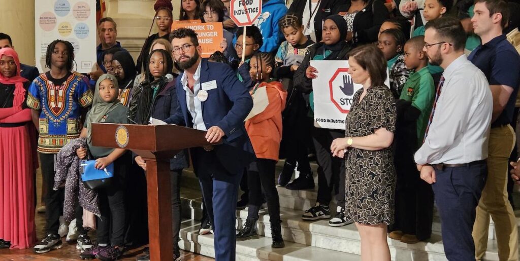 Share Food CEO George Matysik, center, a White man in a blue suit with short brown hair and beard and glasses, stands behind a podium at a protest rally in Harrisburg, PA.