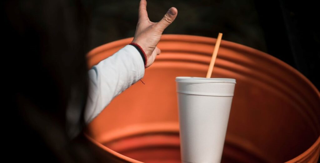A long-haired person in the midst of throwing a foam drinking cup and straw in an orange trash bin