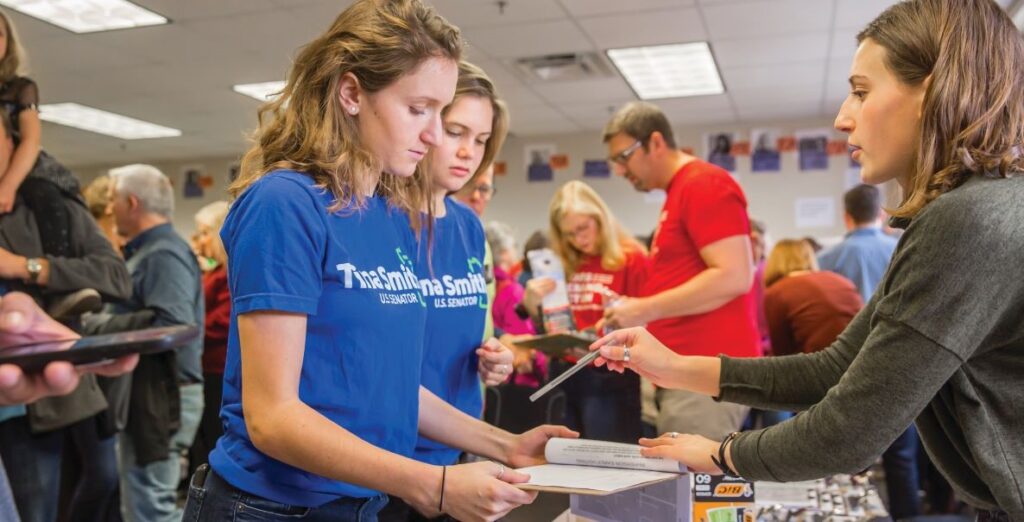 In the foreground, a white young woman in a blue t-shirt with illegible writing holds and looks at a book while facing another young white woman in a grey t-shirt over a long-sleeved grey t-shirt. They are in a room of people who all seem to be concentrating on papers.