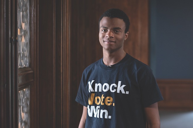 Baxter Montgomery, an African American male college student with short hair wearing a black t-shirt with the words "Knock. Vote. Win." stands in front of a dark wooden wall, looking out with a slight smile.