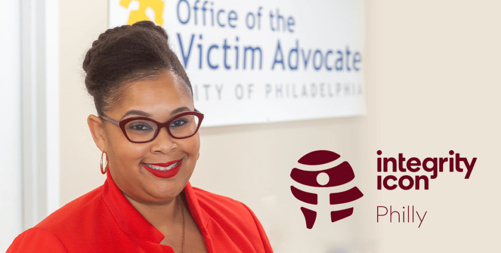 A shoulders-up portrait of Adara Combs, Philadelphia Victim Advocate and 2024 Integrity Icon. Combs is a Black woman with pulled back and braided hair, wearing red-frame glasses, red lipstick, gold hoops and a red top. She is smiling. Behind her is a sign reading "Office of the Victim Advocate CITY OF PHILADELPHIA."