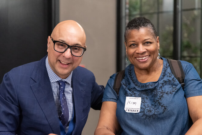 Ali Velshi, a bald Indian American man wearing a navy suit jacket and tie and white and blue shirt, smiles alongside Kim Cooper, a Black woman with short hair wearing an embroidered blue short-sleeve shirt and backpack and also smiling.