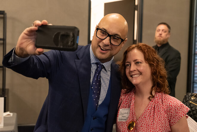 Ali Velshi, a bald Indian American man wearing a navy suit jacket and tie and white and blue shirt, and Jamie Duguay, a white woman with curly ginger hair and a red and white v-neck dress, smile as Velshi take a selfie photo of the two of them.