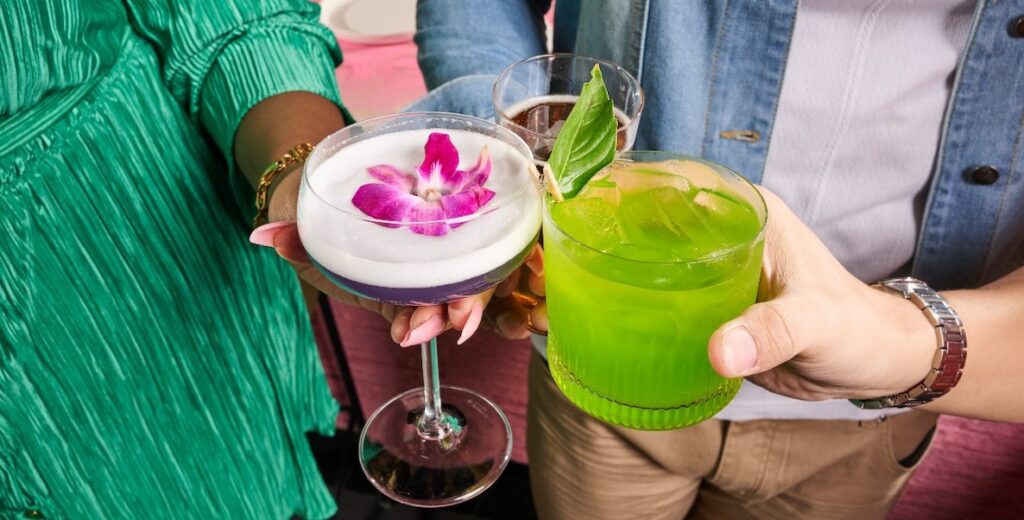 Two hands hold colorful cocktails, one with a flower in the top of a martini glass, another in a rocks glass the color of aloe.