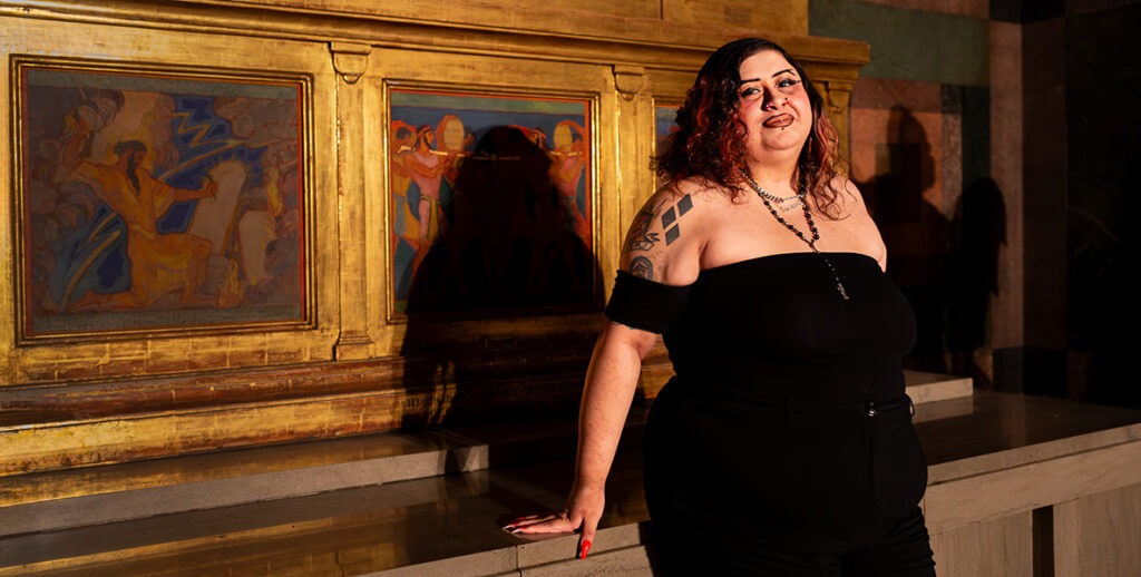 Artist Chela Ixcopal, a Latinx artist with curly shoulder-length orange and black hair, tattoos on one shoulder, a necklace, wearing an off-the-shoulder gown, stands before paintings.