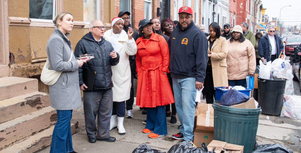 Mayor Cherelle Parker (center, a Black woman in a bright red-orange trench coat, black baseball cap, jeans and orange sneakers) and Carlton Williams (Black man in blue jacket, jeans and red cap) on a "Clean and Green" walking tour in February 2024. A crowd of about 10 people stands behind them on a sidewalk. To one side are rowhomes; to the other are bags, cans and boxes of trash. Photo by Albert Lee.