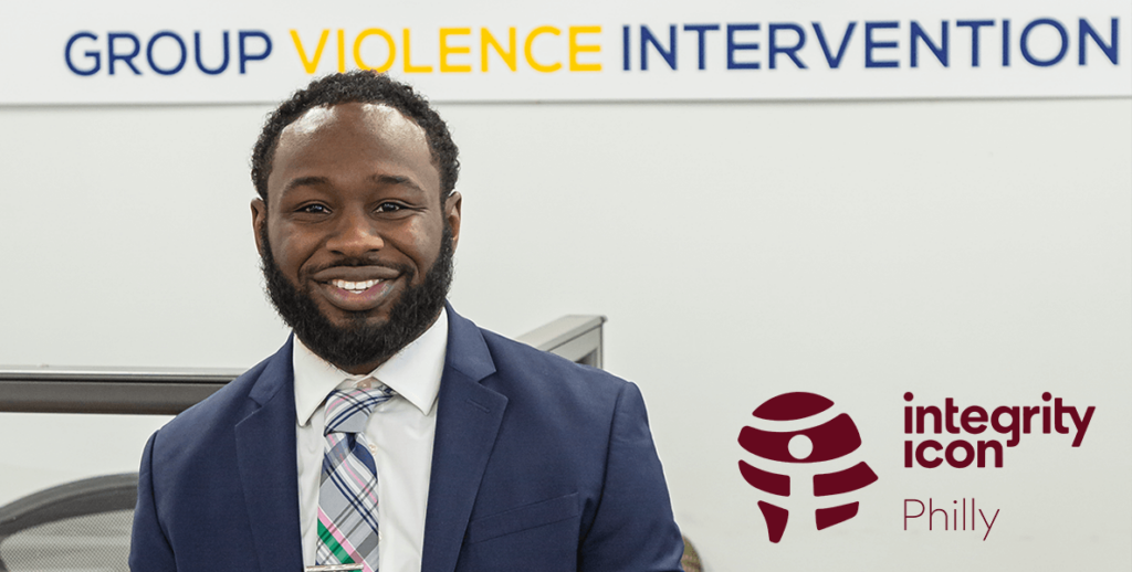 A torso-up portrait of Deion Sumpter, Director of Group Violence Intervention for the City of Philadelphia and 2024 Integrity Icon. Sumpter is a Black man with a close beard and curly hair. He is smiling and wearing a navy suit jacket, white shirt and plaid tie. Behind him is a sign reading "GROUP VIOLENCE INTERVENTION."