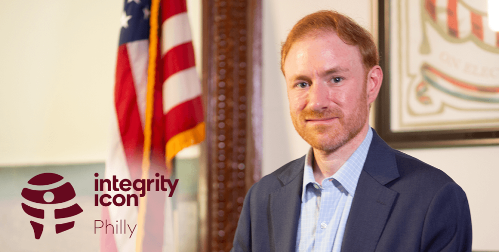 Eric Kapenstein, Philadelphia Deputy City Commissioner and 2024 Integrity Icon. A torso-up portrait of Eric, a White man with ginger hair and beard and blue eyes wearing a light blue shirt and navy suit jacket. Beside him is a U.S. flag.