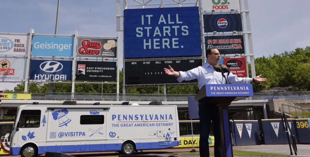 PA Governor Josh Shapiro, a White man with short dark hair, glasses wearing a blue button-down shirt and dark pants, stands before a podium with a sigh reading "PENNSYLVANIA, THE GREAT AMERICAN GETAWAY" with arms outstretched while speaking into microphones. Behind him is a white, blue and red RV with the same slogan and "@VISITPA," and signage from PNC Park, including a large blue sign with white lettering reading "IT ALL STARTS HERE."