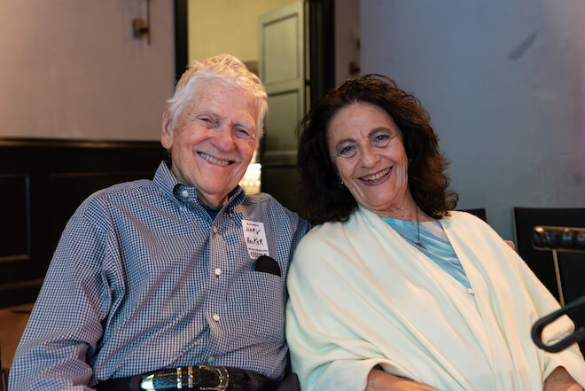 Harv Becker, an older white man with white hair wearing a blue and white gingham buttondown sits with his arm around Joan Becker, an older woman with curly brown hair wearing a blue shirt and cream color wrap.