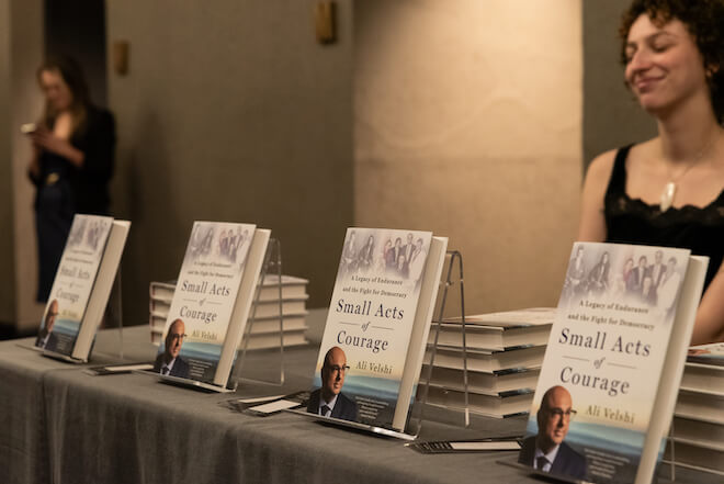 Four hardback books featuring the image of Ali Velshi and the words "Small Acts of Courage" are propped on a table in front of stacks of the same book. Behind the table sits a white woman with curly brown short hair wearing a black tank top.
