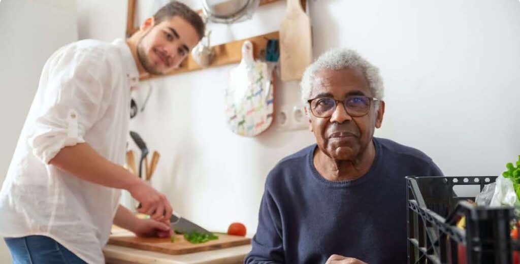 In the foreground, an older Black person with white hair, glasses and a navy blue sweater appears to be seated at a table. To his side is a plastic bin of produce. In the background, a younger person stands to chop a green vegetable on a cutting board. This person is white, with short brown hair, a short beard, white shirt and jeans. Both look toward the camera.