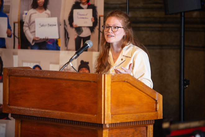 Jessica Bachman, a White woman with long ginger hair pulled back at the top, wears a white jacket and glasses and stands before a wooden podium, speaking into a microphone. Behind her hang blurry photos of people holding white signs with two illegible words on them, at The People’s Budget Roundtable at City Hall. Photo by Steve Weinik.