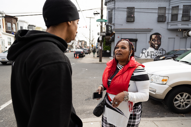 John Solomon, founder of Endangered Kind at N. 24th Street and W. Somerset Street, where he grew up, stands on a street corner speaking with North Philly resident Aisha Best, a Black woman with a red vest, white and black striped sweater, and black and white striped pants and jewelry. Solomon is a Black man with a mustache. He is wearing a black beanie hat, black hoodie.