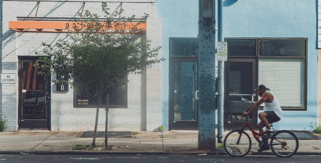 A person wearing a white tank top and shorts rides a bike along a city street, past a blue and a white storefronts. This is 3312 Kensington Avenue (with the orange awning), occupied by Vizionz Gallery, is one of the several properties KCT stewards. Photo by Luis Acosta Studio, 2020.