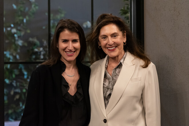 Leah Popowich, a White woman with long brown ahri and a collared black outfit stands alongside Susanna Lachs, an older white woman with long brown hair wearing a cream color suit jacket and patterned blouse. The are smiling and standing before a tall paned window.