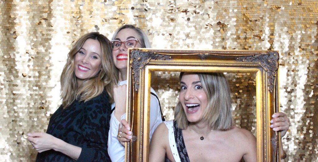 RadGirls founder Leah Kauffman, in frame, with designers Kiley Baun and Betsy Helm in 2019. Pictured are three white women with blonde hair and makeup smiling before a gold-sequined curtain. One of the women is holding a gilded frame around her face.