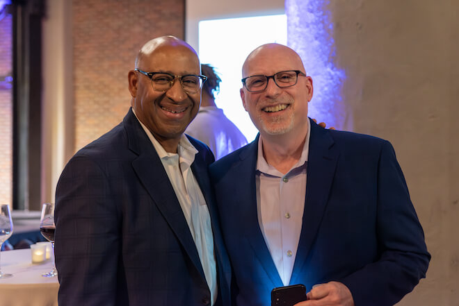 Michael Nutter, a bald Black man in glasses, a dark suit jacket and white shirt, smiles as he stands next to Larry Platt, a bald white man with glasses, a navy suit jacket and lavender button down shirt, also smiling and holding a phone.