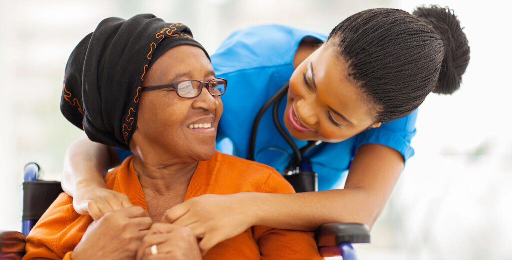 An older African American woman with a black and orange head wrap, glasses and orange top sits in a wheelchair and holds the outstretched hands of an African American nurse wearing scrubs, a stethoscope, and small braids pulled into a bun leans over her from behind. Both smile.