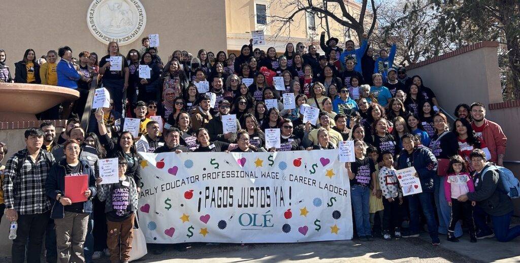 A large group of people stands on steps outside an official building in New Mexico. They are carrying signs in support of universal childcare from Olé, an organization that advocates for free childcare.