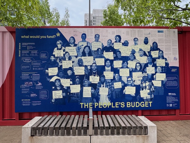 Outside The People's Budget Office is a large photo-style blue and white mural of people holding signs. Around them is small text. The mural reads in larger print: "what woudl you fund?*" and "THE PEOPLE'S BUDGET." It hangs on a converted shipping contained that's painted red. Behind it are trees and the top of a building. In front of it is a concrete and wooden bench and brick ground. Photo by Phoebe Bachman.