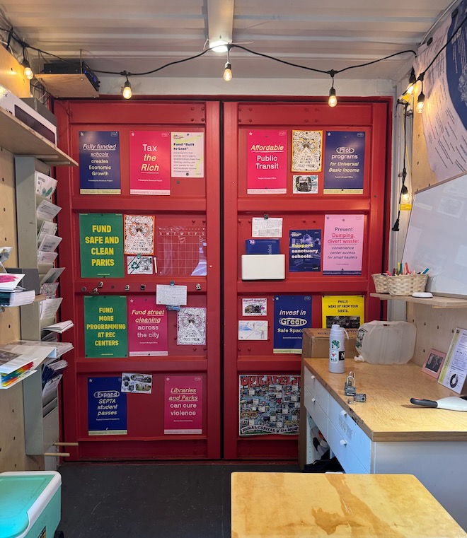 Inside The People's Budget Office, a red wall with colorful flyers hanging on it. This wall is inside a modified shipping container, with white walls, a strand of iridescent bulbs, two desks and a wall shelf of papers. Photo by Phoebe Bachman.