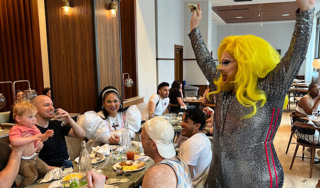 A drag queen in a bright long yellow wig and silvery bodysuit stands by a table of people having brunch.