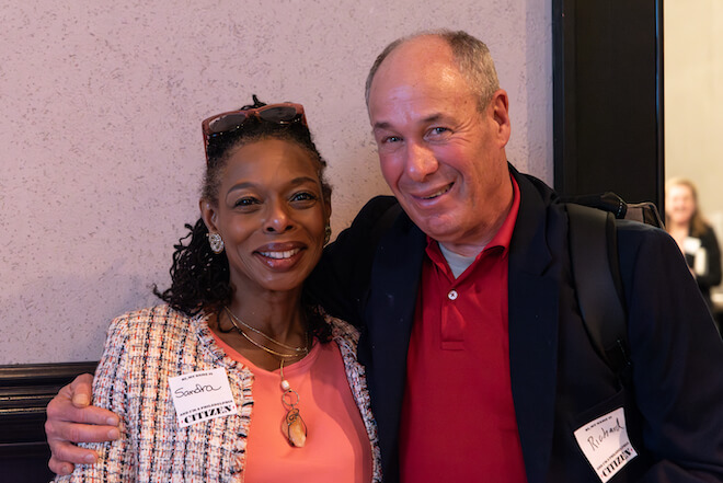 Sandra Dungee Glenn, a Black woman with curly shoulder length black hair, glasses atop her head, a peach-color top, long gold necklace and Chanel style suit jacket stands beside Richard Binswanger, a partially bald white man in a red polo shirt and navy suit jacket. Richard has his arm around Sandra. Both are smiling.