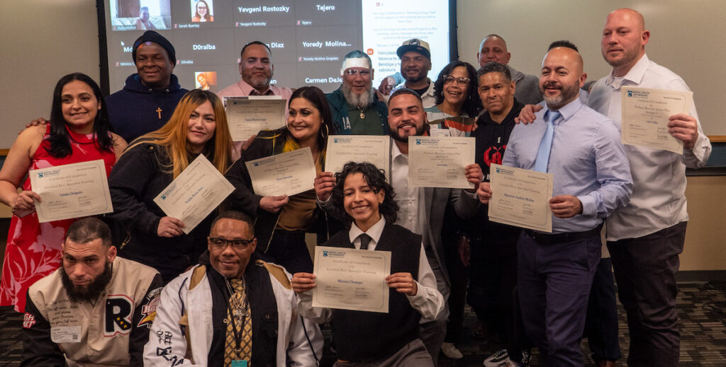 A group of 15 people pose for a photo holding certificates at a Mental Health Partnership graduation ceremony for Spanish-speaking Certified Peer Specialists.
