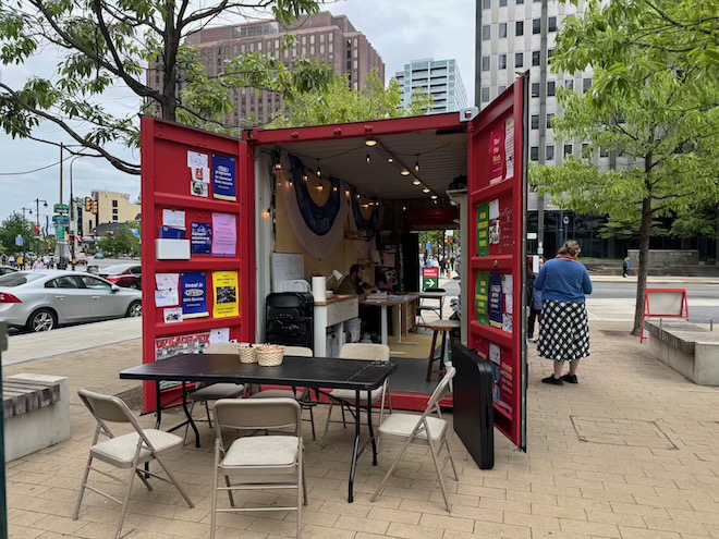 Outside in a city setting, a converted shipping container painted red stands open at both ends, revealing flyers posted to walls, desks and lights. At the closer end of the container are six folding chairs and a folding table. Outside the container are trees and treetops, and a city view and one person wearing a long dotted skirt and blue sweater. This is The People's Budget Office in LOVE Park.