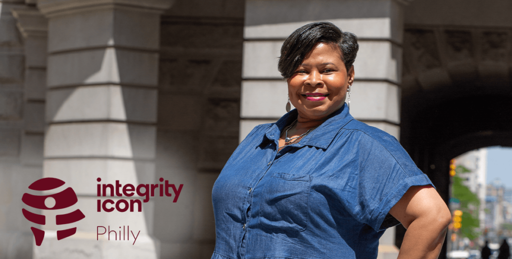 Tracey T. Williams, Deputy Commissioner for the Department of Records and 2024 Integrity Icon. Her portrait is taken outside City Hall. She is a Black woman with short hair wearing a denim blue top, earrings, a necklace and red lipstick.