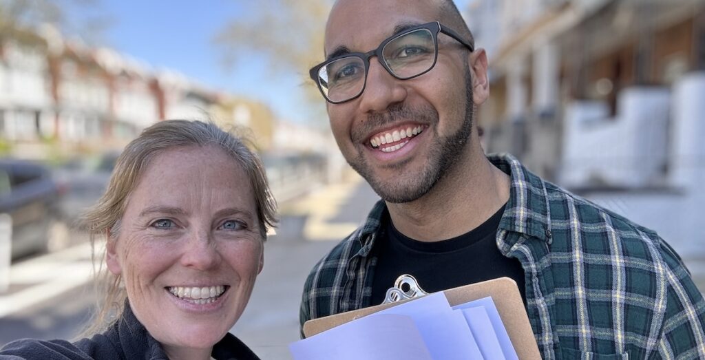A close-up photo of two smiling people's heads, one white woman with ginger hair pulled back and a light-skinned African American man with a light beard and glasses holding a clipboard and white papers.