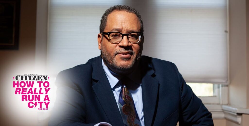 Michael Eric Dyson, a Black man with short, curly hair, black-and-clear-rimmed glasses, a short grey beard looks forward. He appears to be seated and is wearing a navy blue suit jacket, light blue collared shirt, and multicolored tie.