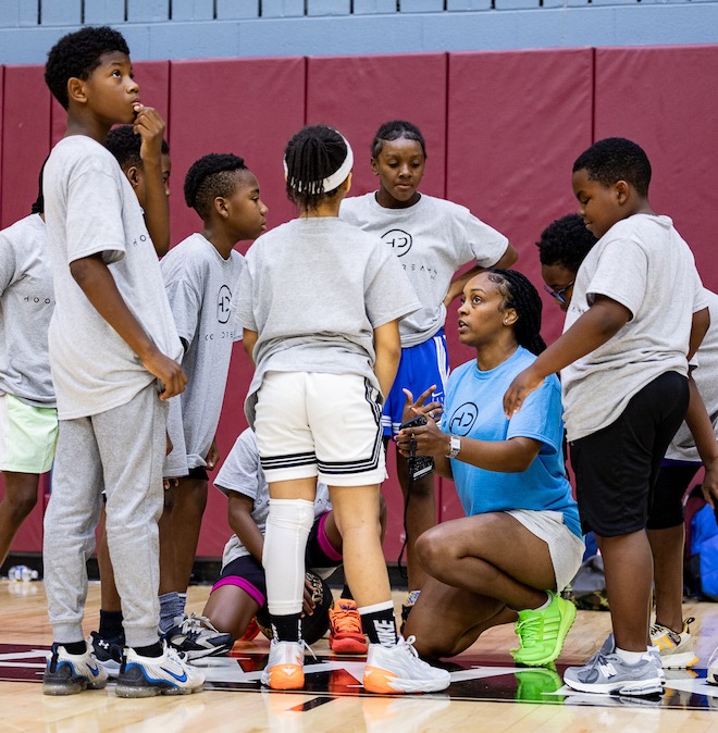 A group of about seven Black children wearing shorts, t-shirts, sweats and sweatpants, approximately ages 10-12, surround Nadia Bosket, a Black woman wearing a blue t-shirt, white shorts and neon green sneakers, partially kneeling on a basketball court and offering them instructions.