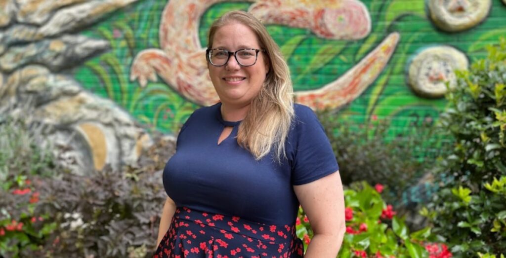 Jacquelilne "Jax" Cusack, a white women with armpit-length blond hair and glasses, wears a navy blue short-sleeved shirt and high-waisted navy and red skirt. She smiles and stands before plants and a brick wall painted with art that's hard to identify.