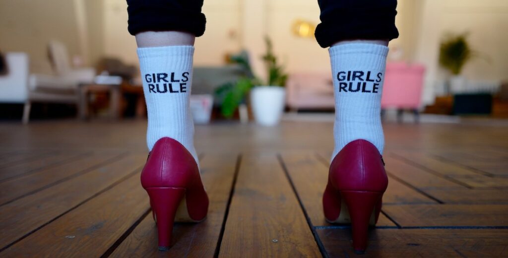 A view of the someone's calves, wearing cuffed black pants and sweatsocks that say "GIRLS RULE" on the back, and bright pink heels.