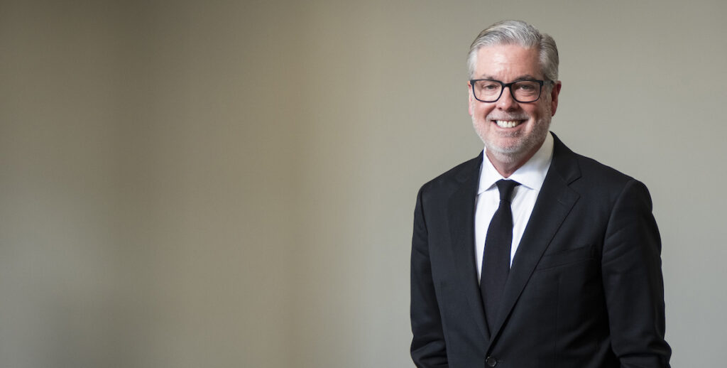 John Fry, a White man with short grey and white hair and glasses, smiles. He is wearing a back suit jacket and tie and white shirt.