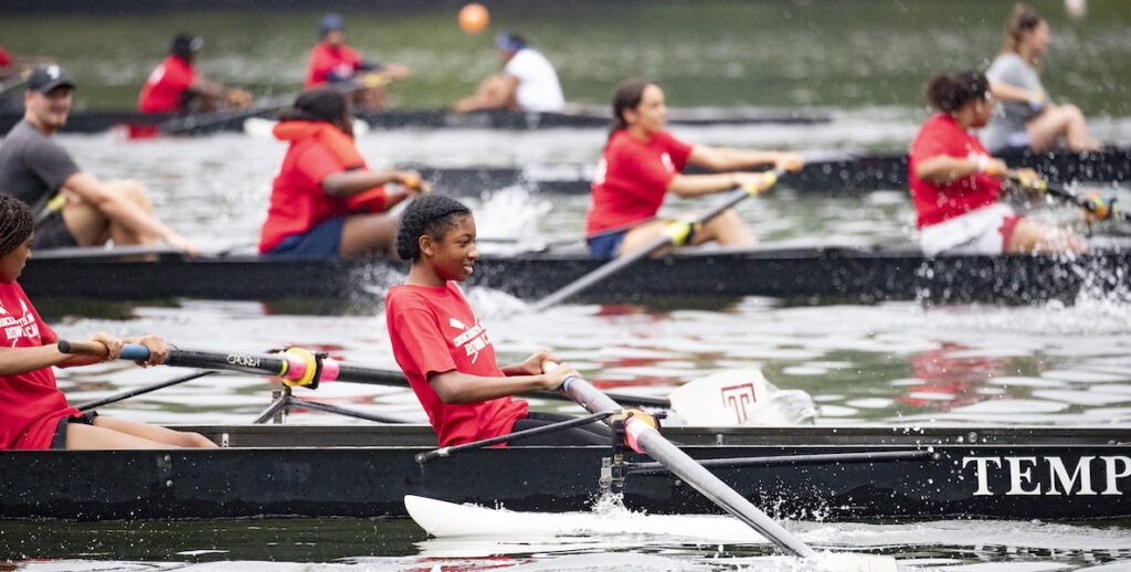 Middle school students in bright red t-shirts row in Temple boats along the Schuylkill River.