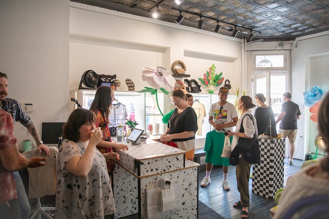 Inside a small corner clothing boutique with tin ceilings and white walls, a group of people line up to pay at a white counter splashed with black paint.