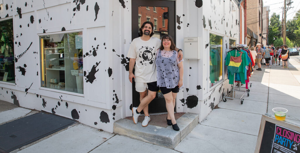 Two people, a man with a dark beard and almost shoulder-length black hair, wearing a t-shirt with a roaring cat at the center and black and white shorts, stands with his arm around a shorter woman wearing a sheer, white top splattered with black paint and short black bottoms. They are standing in front of a corner shop painted white with black splatters, where a sign on the sidewalk says "closing party" and a rack of colorful clothing stands alongside one of the shop's outdoor walls along the sidewalk.